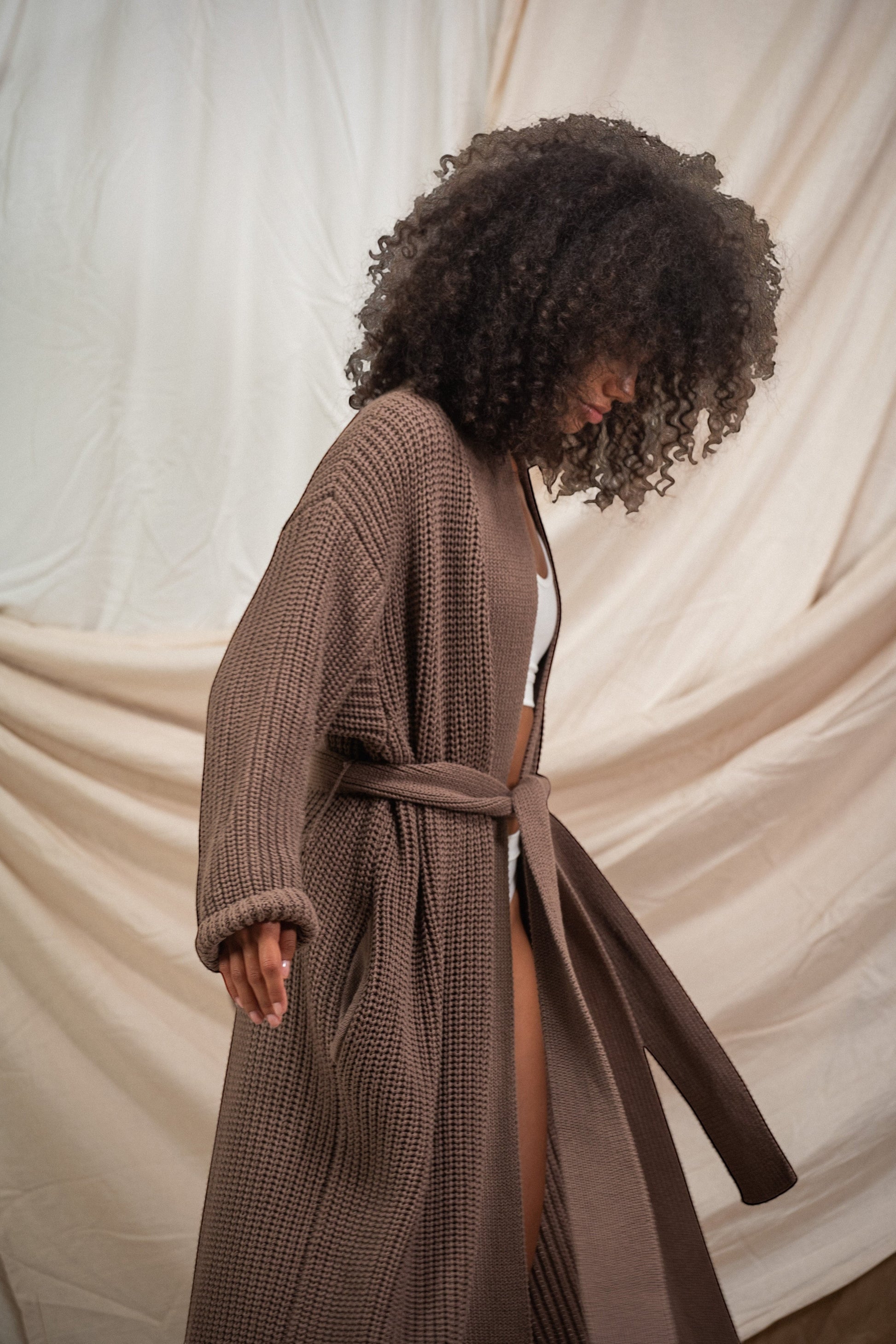 AMI CHÉRIE DESIGN STUDIO longline brown knit robe with belt and pockets, worn by a model with curly hair.