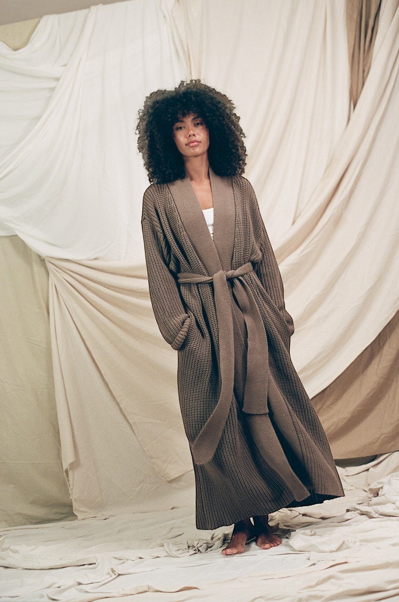 AMI CHÉRIE DESIGN STUDIO longline brown knit robe with belt, V-neck, and pockets, worn by a model with curly hair.